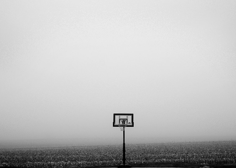 Outdoor basketball backboard and hoop with a pole in the ground looking small in the center of the image set in front of a vast field and open sky.
