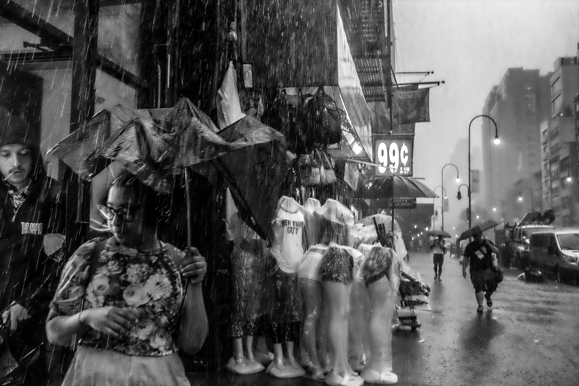 A woman with dark-framed glasses and a flowered top is caught in a rainstorm on W 14th Street in New York City with only a crumpled umbrella to try to stay dry. A group of headless full-body and torso mannequins are close by and covered in plastic.