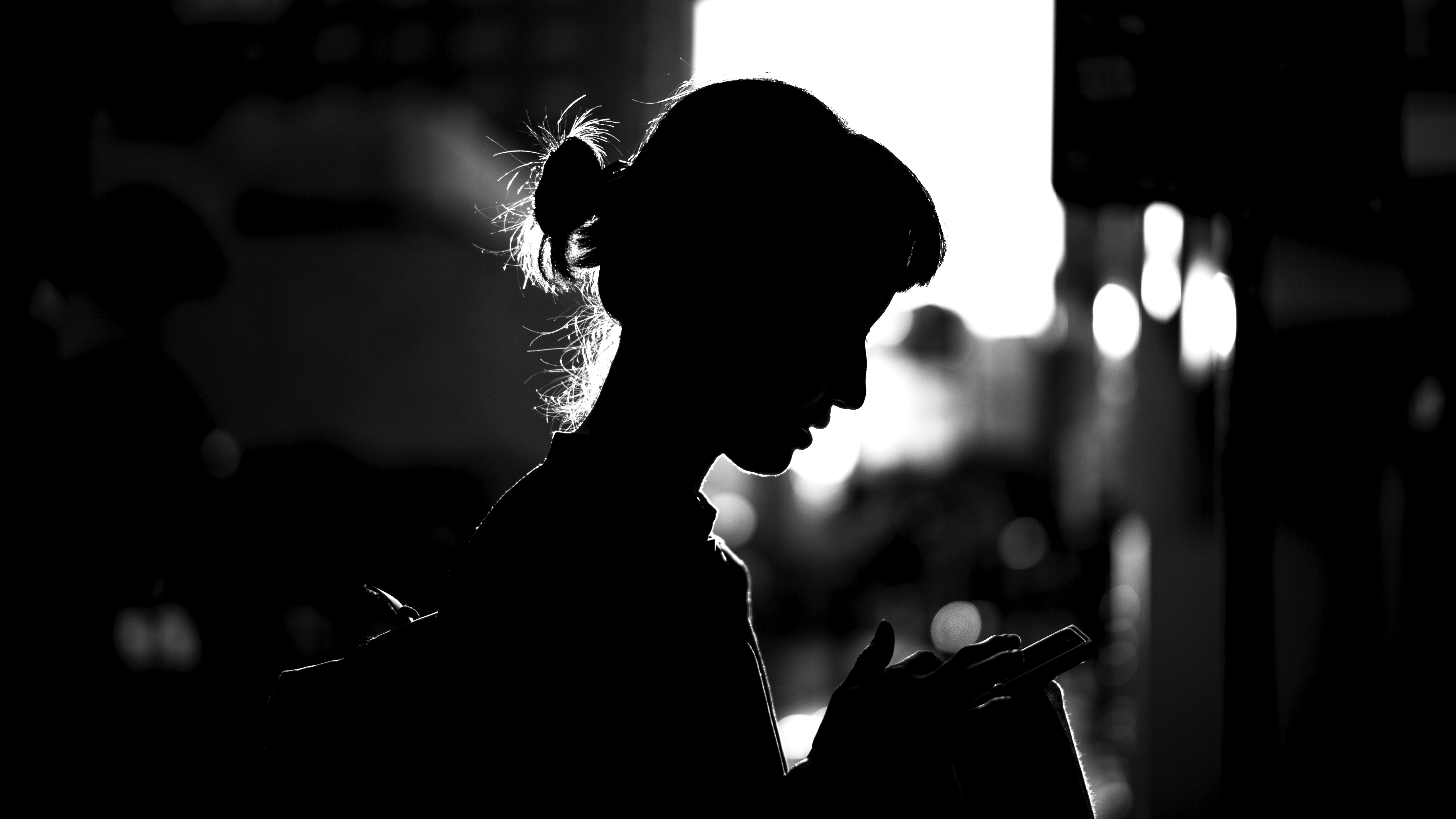 A young woman in silhouette with a messy bun and sporting a backpack stands looking down at the device she holds. The buildings in the background are blurred.