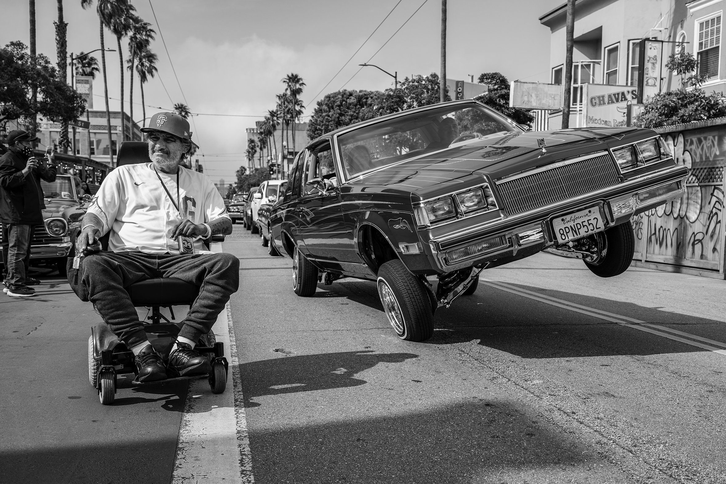A man in a San Francisco cap sits in a power chair along the edge-line marking of a street and very close to a car balanced on its right two wheels during a Cinco de Mayo celebration organized by the San Francisco Lowrider Council.
