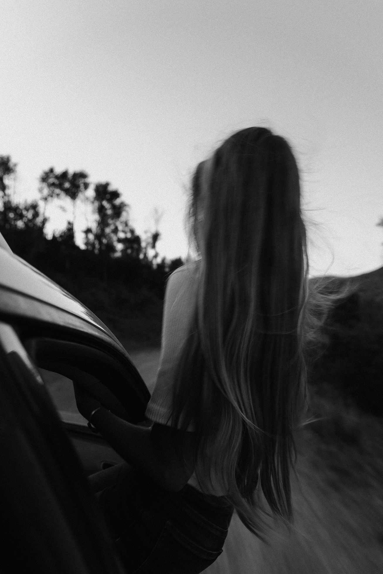 A girl with waist-length hair is seen from the back as she sits on the frame of the front passenger’s open car window with only her legs inside.