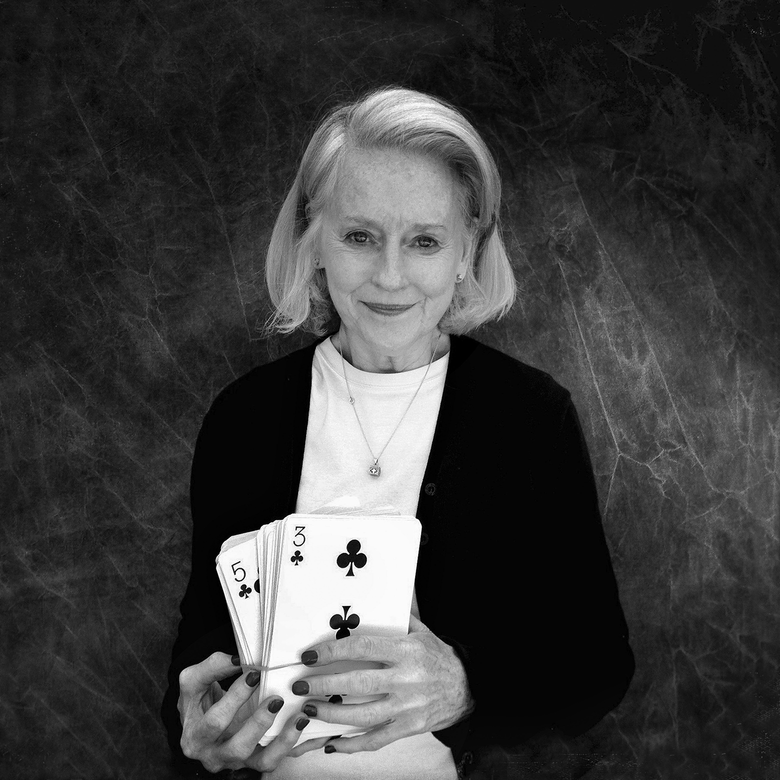 An older woman with a closed-mouth smile in a white T-shirt and black sweater holds up an outsize deck of playing cards with a rubber band around it and the three and five of clubs are visible.