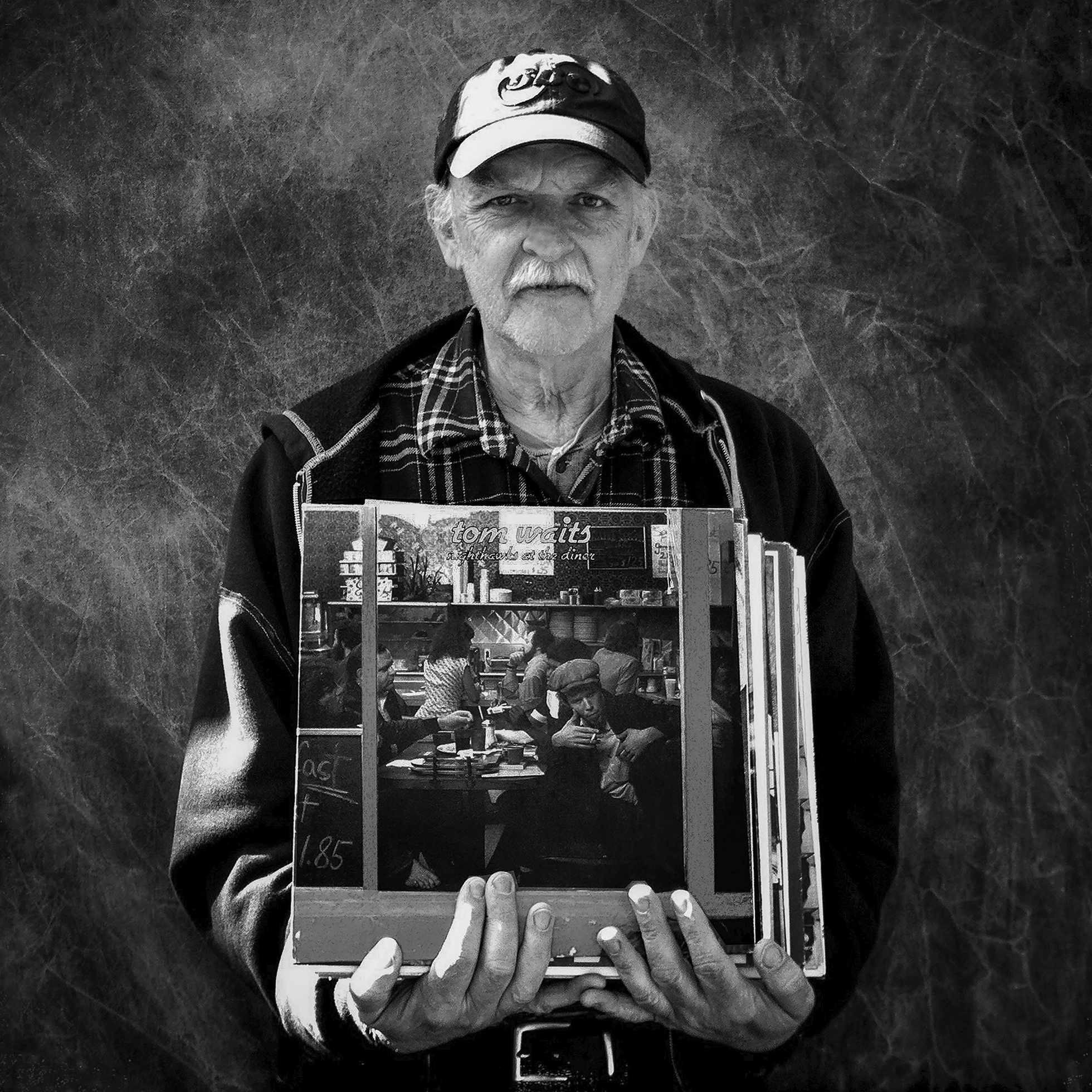 An older man in a cap and jacket holds a stack of albums against his chest presenting them to the camera. The album cover that is visible is Nighthawks at the Diner by Tom Waits.