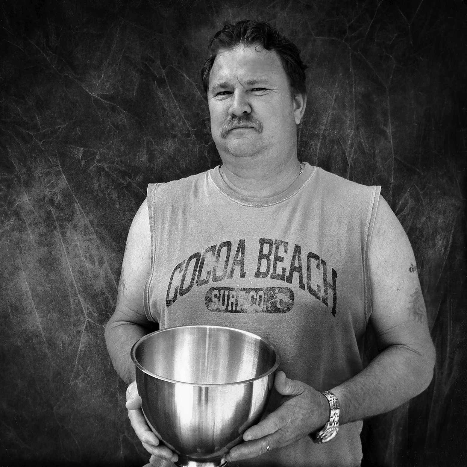 A man with a moustache wearing a sleeveless T-shirt that reads “Cocoa Beach Surf Co.” holds a metal mixing bowl.