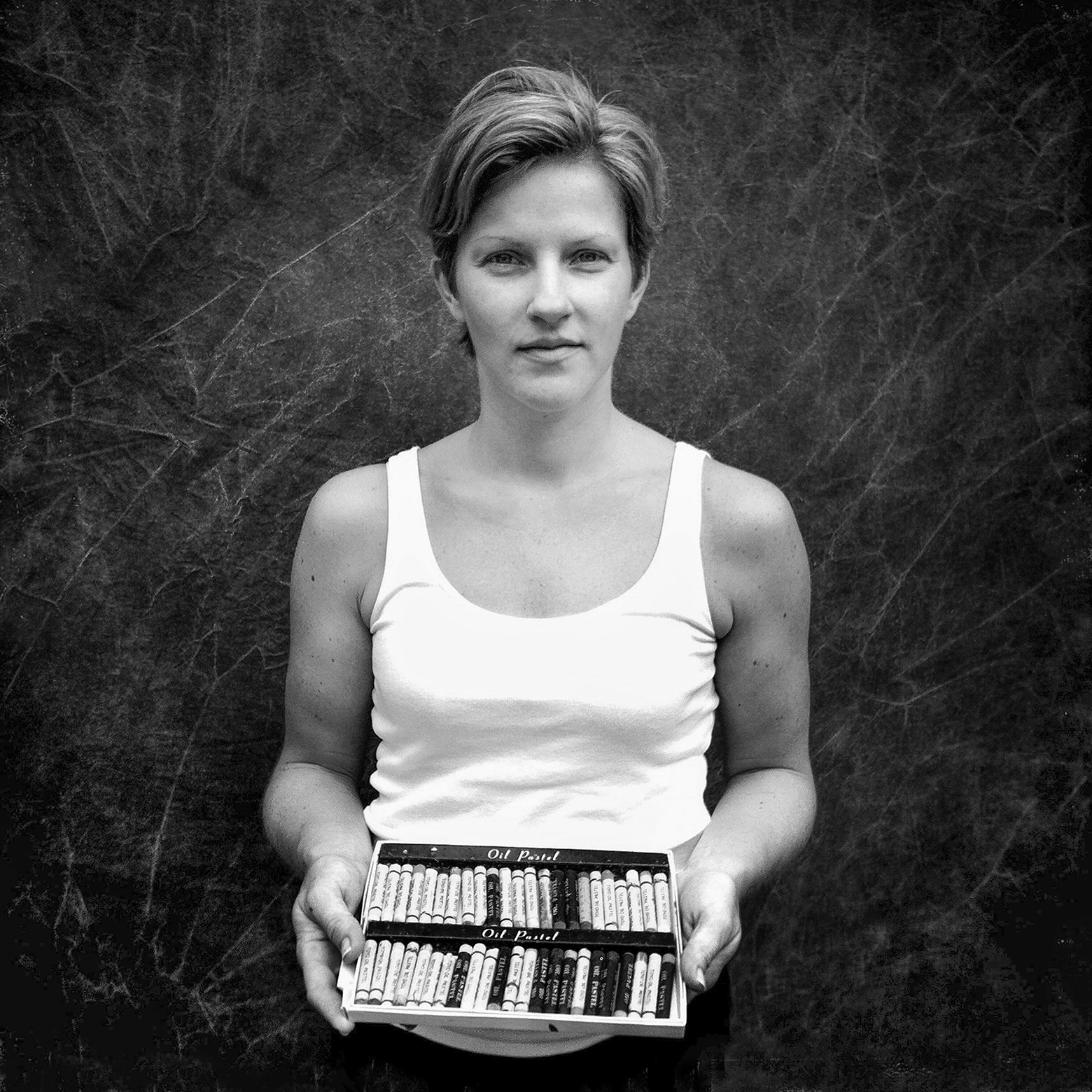 A woman with short hair in a white tank top holds an open box with two rows of drawing oil pastels.