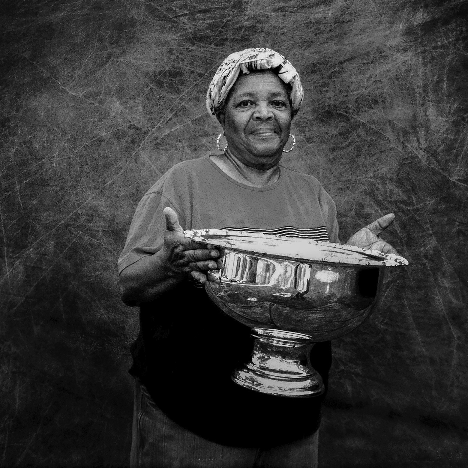 A woman wearing a T-shirt and a headscarf and hoop earrings holds up a large, footed, metal punch bowl.