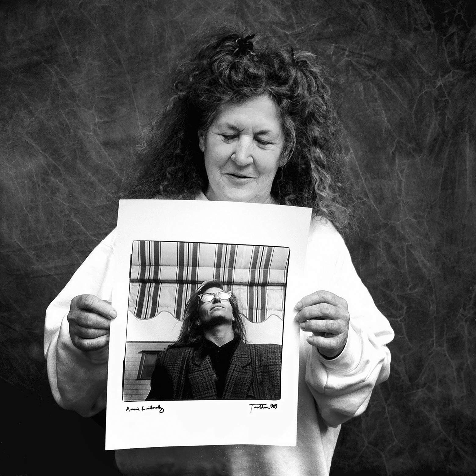 A woman wearing a white sweatshirt with thick, curly, long hair in clips slightly smiles with her eyes closed as she holds up a photograph of Annie Leibovitz in a plaid jacket with shoulder pads taken in front of the Baker Gallery by John Trotter in 1985.