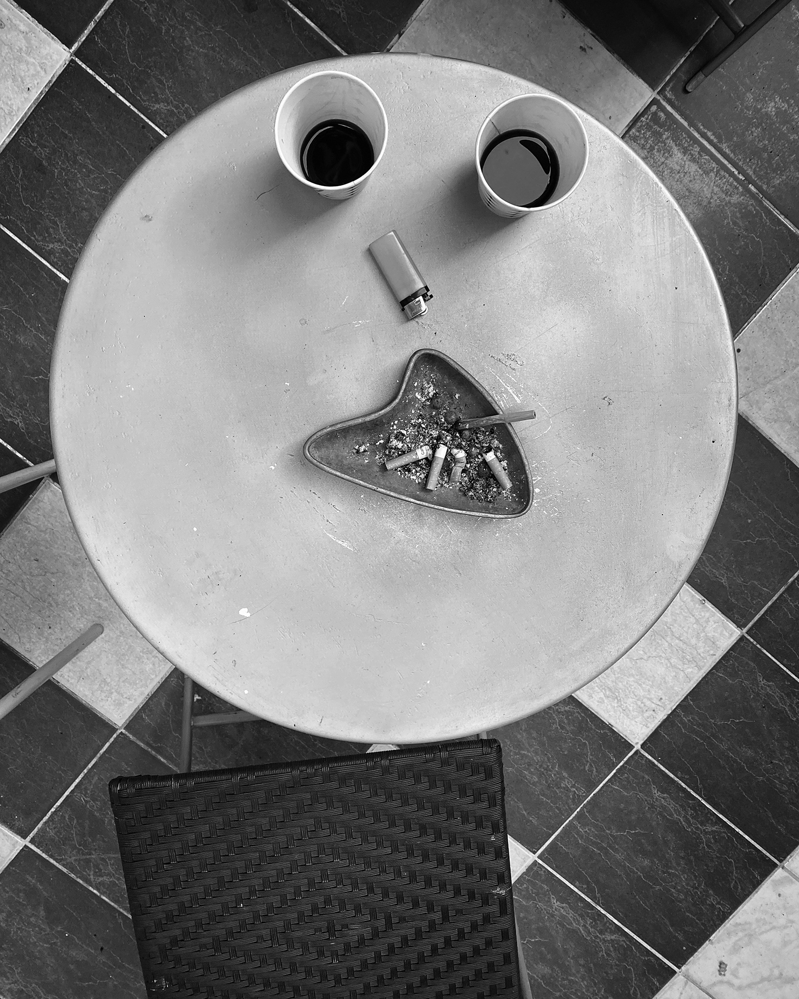 View from above of a round white table against a tiled floor of two contrasting colors. On the table are two disposable coffee cups, a lighter, and an ashtray.