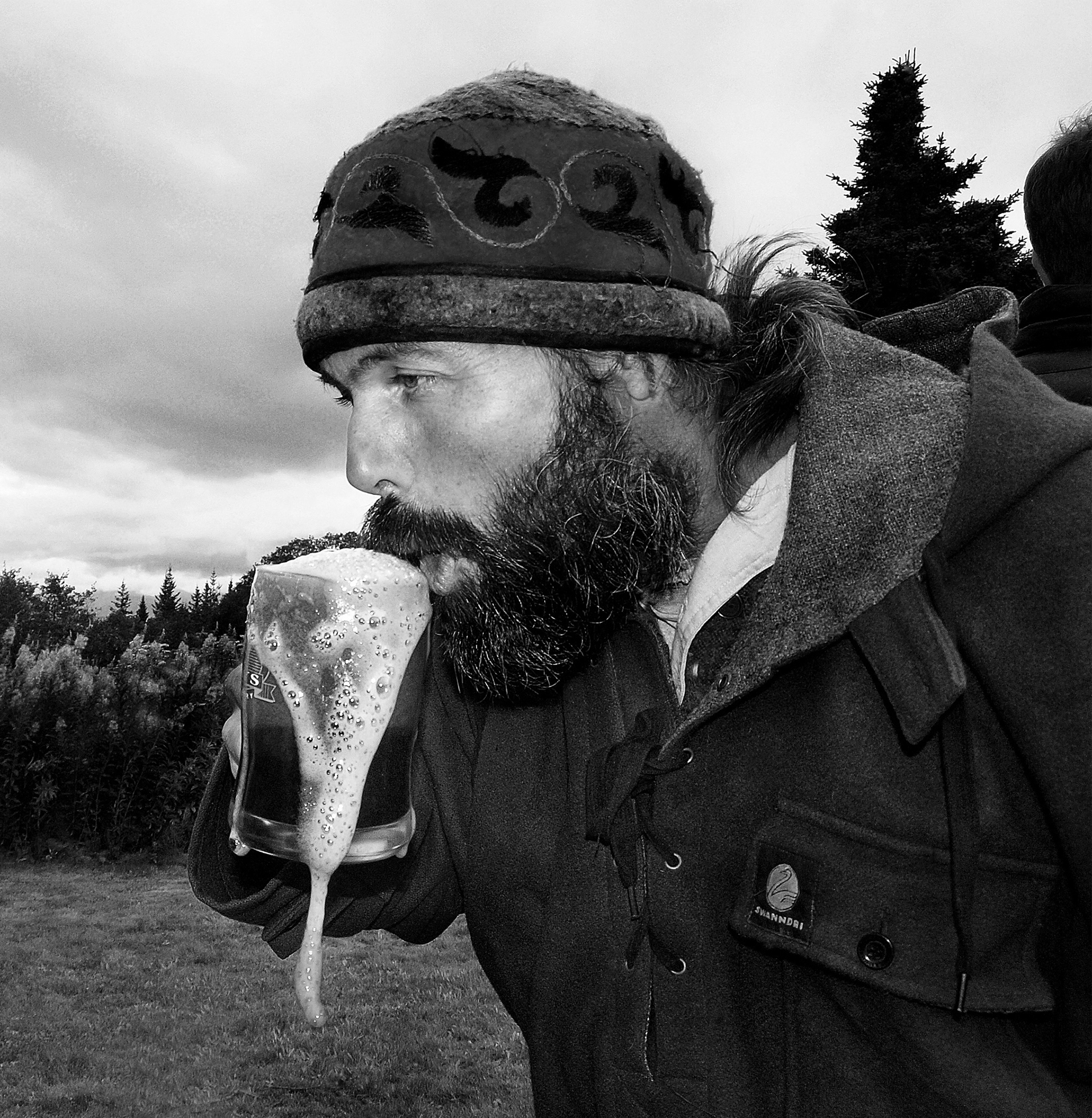 A man with a full beard and mustache wearing a hat and coat stands in a field on a cloudy day and drinks from a glass overflowing with beer.