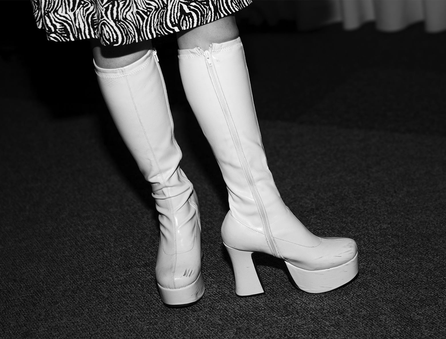 Close-up of a woman standing on carpeting from the bottom of her patterned skirt down to her slightly scuffed white leather platform boots.