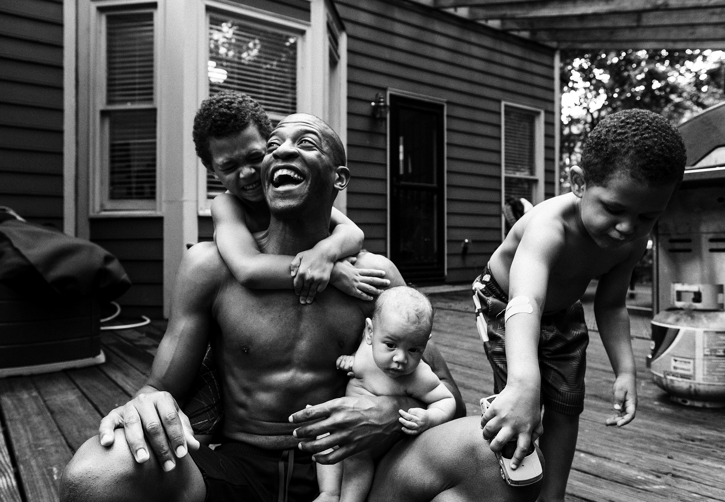 A father and his three sons are outside on the back deck. The father laughs and smiles as he sits and holds the baby while one son has his arms around his father’s shoulders from behind and another son runs a toy car along his father’s knee.