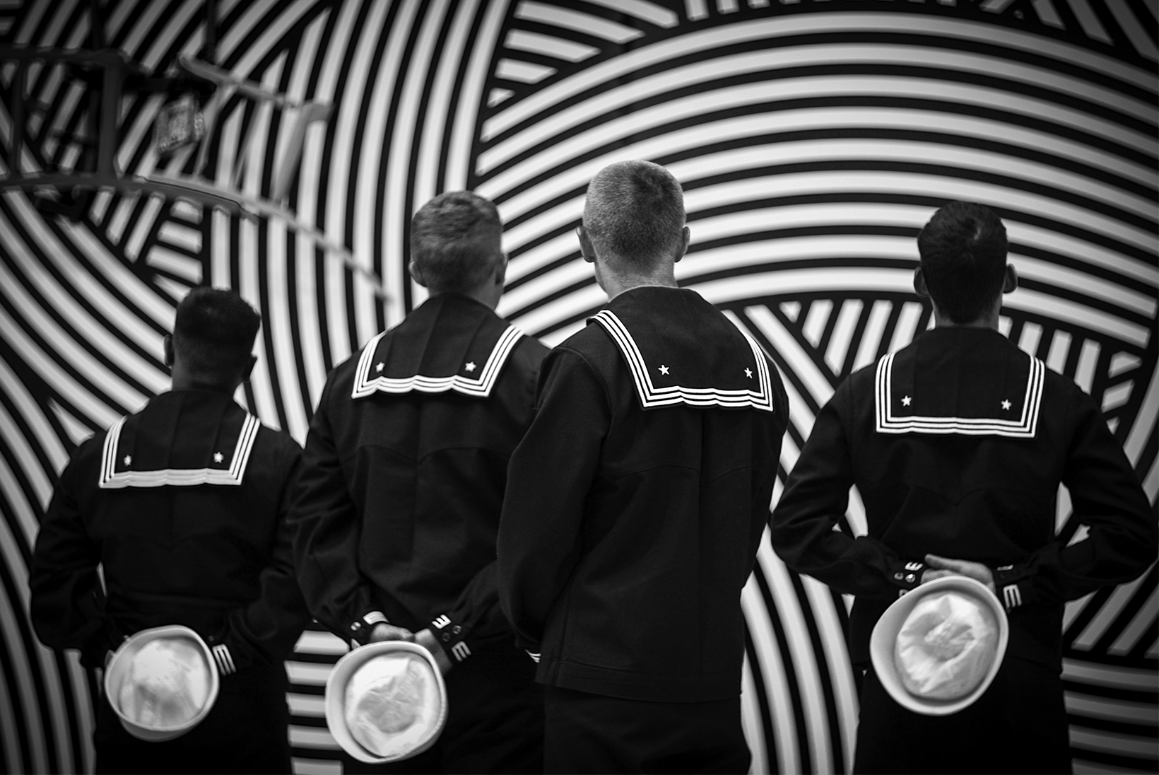 Four sailors in service dress blues seen from behind. Three have their hands behind their backs holding “Dixie cup” hats. They are in front of an art installation with curved, haphazard stripes and three interlocking chairs hanging from the top left.
