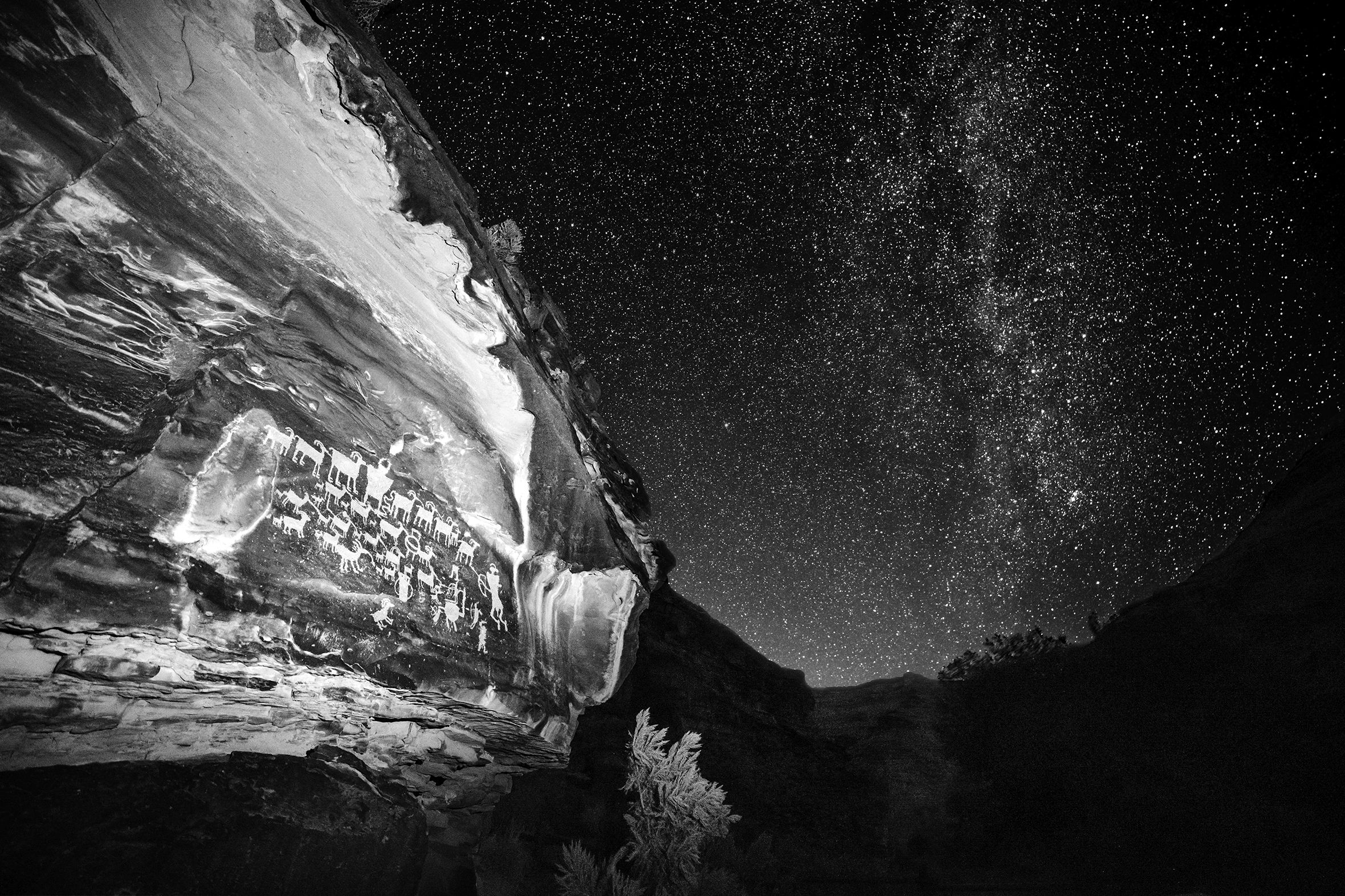 The photo titled “Ancient Nights: Night Hunt” shows Anasazi petroglyphs on the side of a cliff in the foreground against a starry night sky. The petroglyphs are of hunters with bows and arrows surrounded by a herd of large animals.