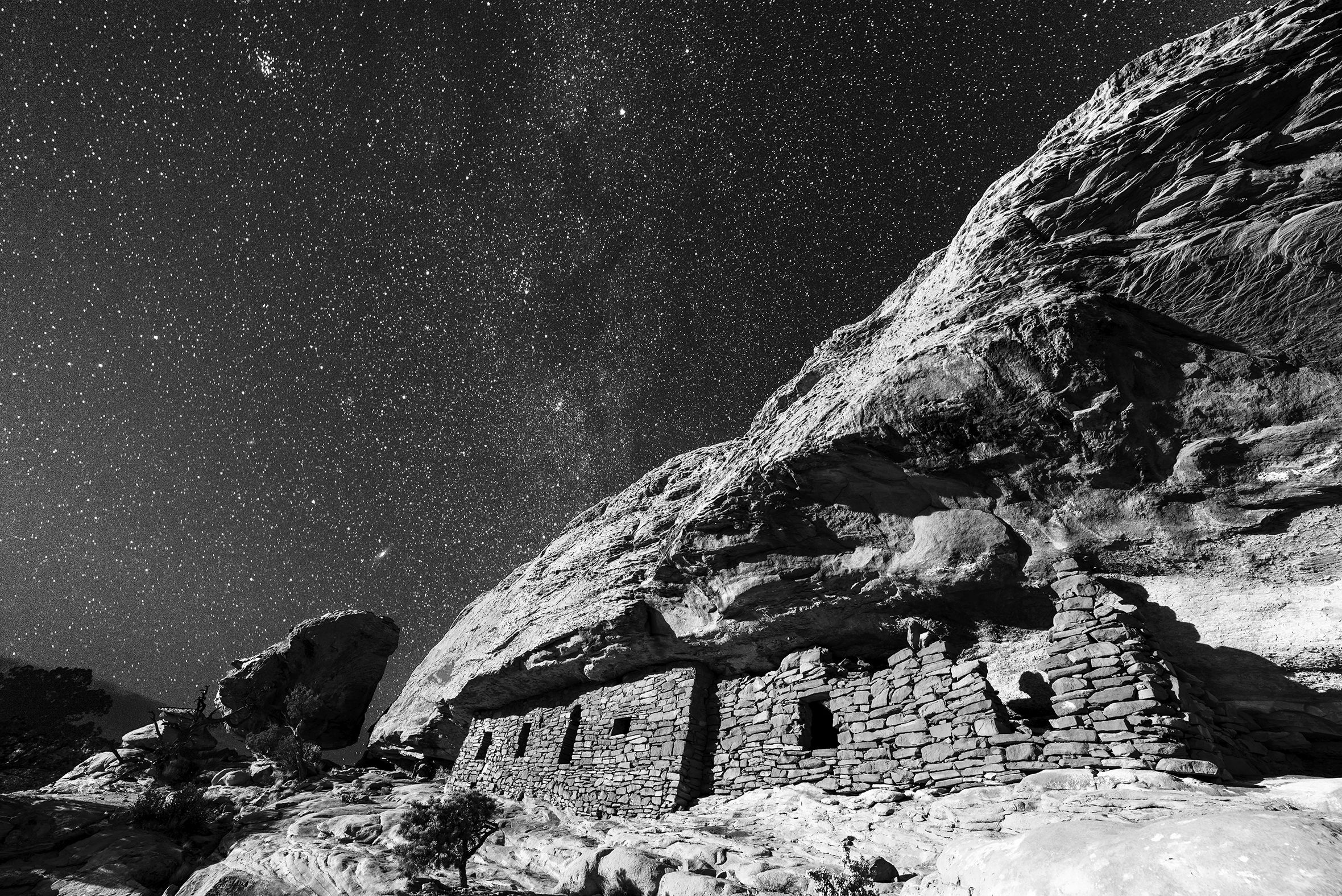 The photo titled “Ancient Nights: The Citadel” shows Anasazi ruins on a starry night. The ruins look like a dwelling of layered stones with several windows built into the side of a cliff under an overhang.