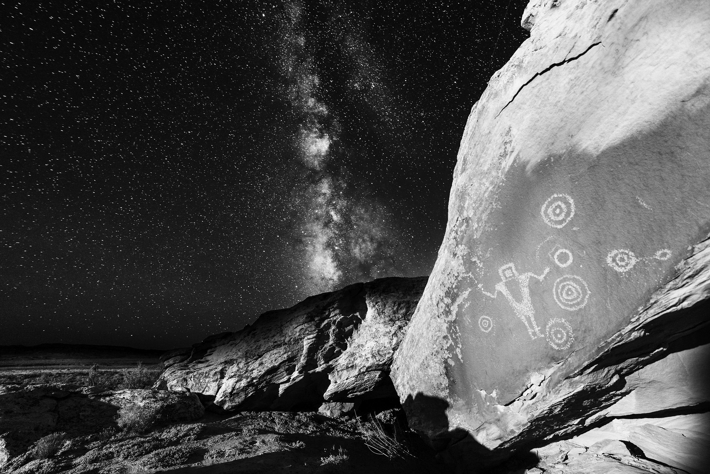 “Ancient Nights: Juggling Fire” shows Anasazi petroglyphs in the foreground against a night sky with a burst of stars. The petroglyphs are of a person with a square head standing with outstretched arms and concentric circles on both sides.